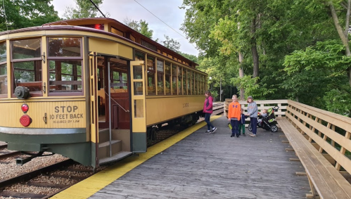 A vintage streetcar stops at a wooden platform surrounded by trees, with people and a stroller nearby.