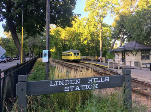 A yellow tram approaches Linden Hills Station, surrounded by trees and a quaint building in the background.