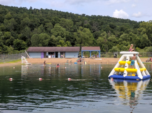 A lake scene with children playing on an inflatable structure, surrounded by trees and a sandy beach area.
