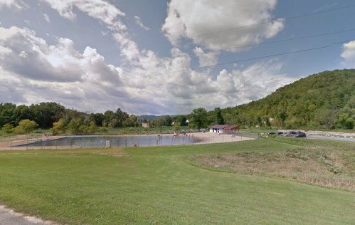 A scenic view of a swimming pool surrounded by green hills and a cloudy sky, with people enjoying the water.