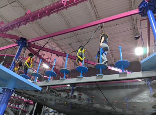 Two people navigate a high ropes course with colorful platforms and safety harnesses in an indoor adventure park.