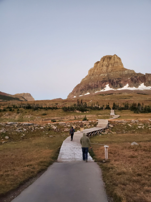 Two people walk along a wooden path through a grassy landscape, with a mountain in the background under a clear sky.