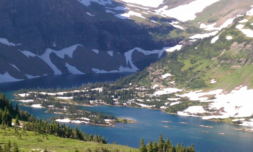 A scenic view of a mountain valley with a blue lake, surrounded by green trees and snow-capped peaks.