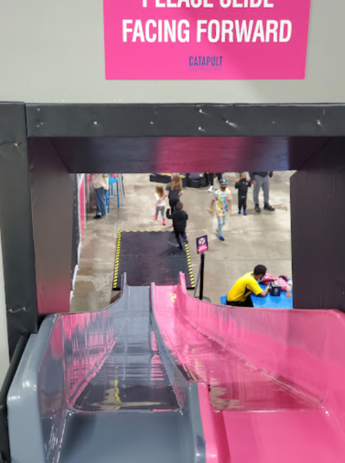 View from the top of a colorful slide, with a sign instructing to slide facing forward and children playing below.