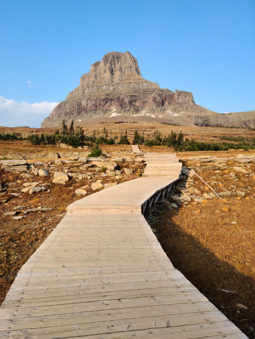 A wooden pathway leads through a rocky landscape towards a towering mountain under a clear blue sky.