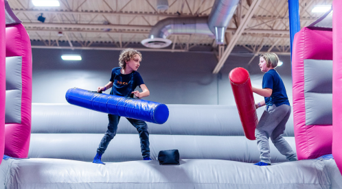 Two kids on a bounce house, playfully battling with foam sticks, one blue and one red, in a fun, energetic setting.