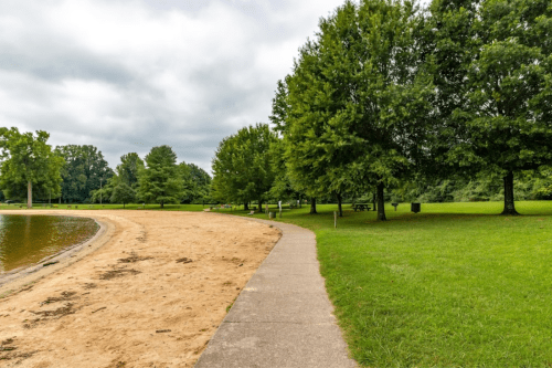 A sandy path curves along a lake, bordered by green grass and trees under a cloudy sky.