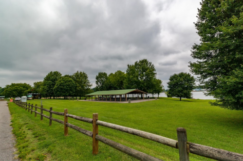 A grassy park with a covered pavilion, trees, and a cloudy sky near a lake, bordered by a wooden fence.