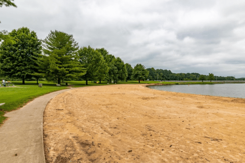 A sandy beach area beside a calm lake, surrounded by green trees under a cloudy sky.