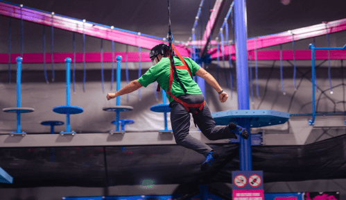 A person in a green shirt suspended in mid-air on a ropes course, wearing a harness and helmet.