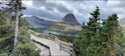 A panoramic view of a mountain landscape with a wooden walkway, surrounded by trees and a lake under a cloudy sky.