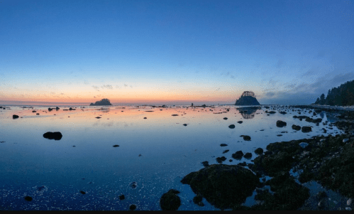 Panoramic view of a calm ocean at sunset, with rocky shores and silhouettes of distant islands under a colorful sky.