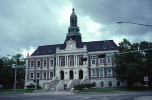 Historic building with a clock tower, surrounded by trees, under a cloudy sky. Traffic lights in the foreground.