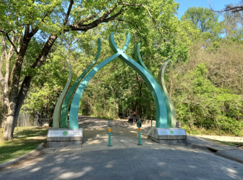 A green archway with curved designs marks the entrance to a park, surrounded by trees and a pathway.