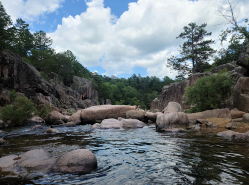 A serene river flows between rocky banks, surrounded by lush greenery and a partly cloudy sky.