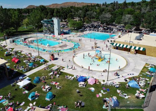 Aerial view of a busy public pool area with multiple pools, sunbathers, and colorful umbrellas on a sunny day.