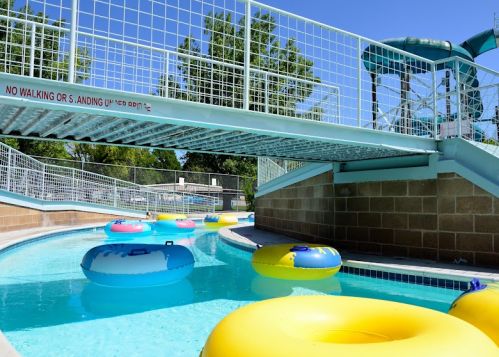 A lazy river with colorful inner tubes, surrounded by a pool area and a bridge overhead under a clear blue sky.