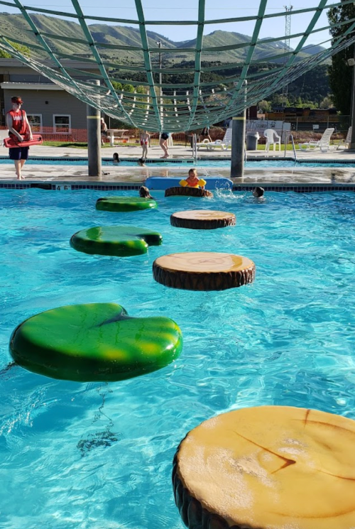 Colorful floating logs in a pool with children playing nearby and a lifeguard watching from the side.