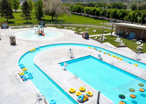 Aerial view of a bright outdoor pool area with slides, floats, and lounge chairs surrounded by green trees.