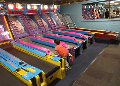 A child in an orange shirt plays at colorful arcade skee-ball machines in a lively game room.