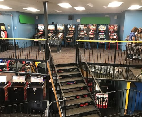 A view of an arcade with pinball machines and video game cabinets, featuring a staircase in the foreground.