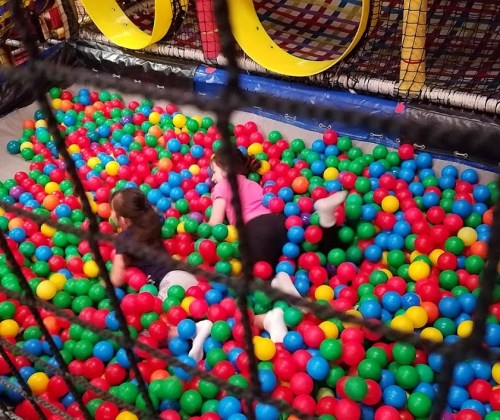 Two children play in a colorful ball pit filled with red, green, and blue balls, surrounded by a netted enclosure.