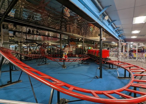A colorful indoor roller coaster with red tracks and empty cars, set in a brightly lit amusement area.