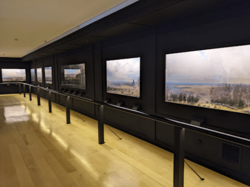 A gallery interior featuring multiple landscape paintings displayed on dark walls with wooden flooring.