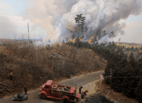 A vintage fire truck near a road, with firefighters battling a large forest fire and smoke billowing from the trees.