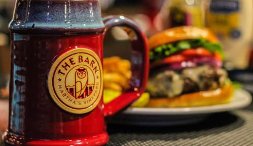 A red mug with "The Barn" logo beside a plate of a burger, fries, and condiments in a cozy dining setting.