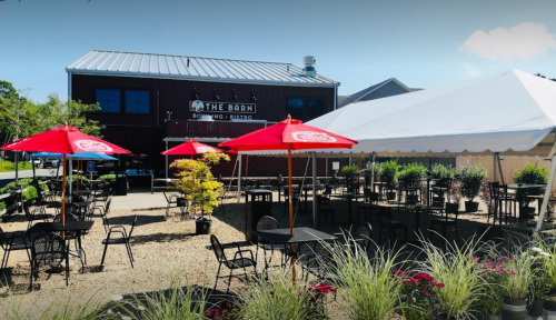 Outdoor seating area with red umbrellas at The Barn, a rustic building, surrounded by greenery and gravel.