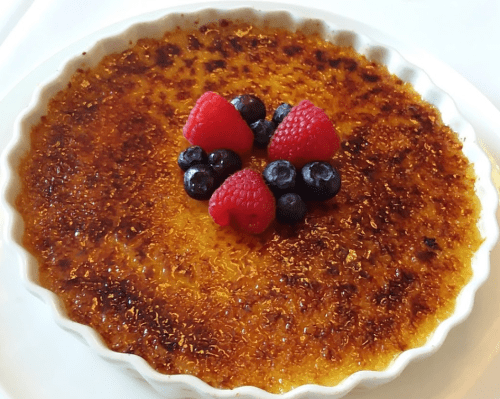 A creamy crème brûlée topped with fresh raspberries and blueberries in a white dish.