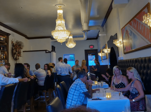 A lively restaurant scene with diners at tables, chandeliers overhead, and staff serving in a warm, inviting atmosphere.