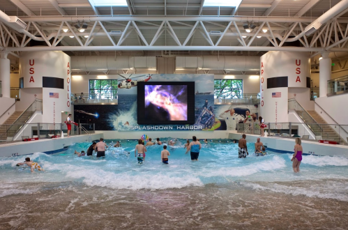 A lively indoor water park with children playing in a wave pool and a large screen displaying colorful visuals.