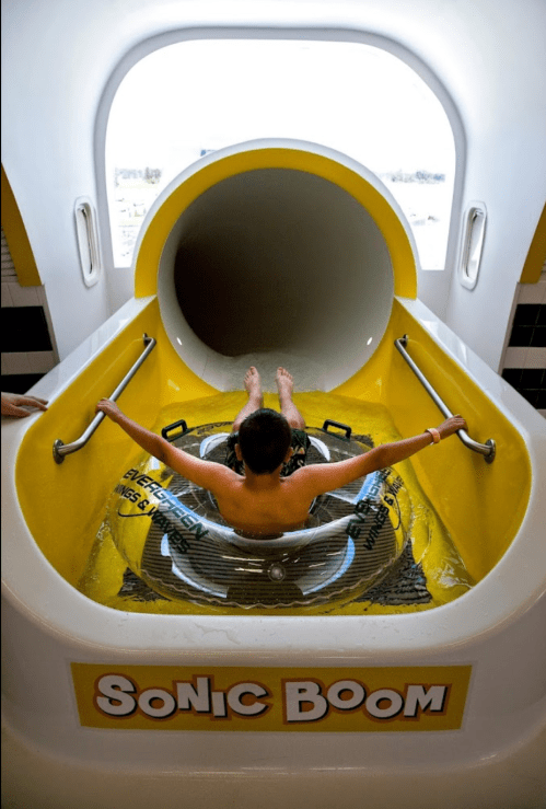 A child sits in a circular raft at the entrance of a water slide labeled "Sonic Boom," ready to slide down.