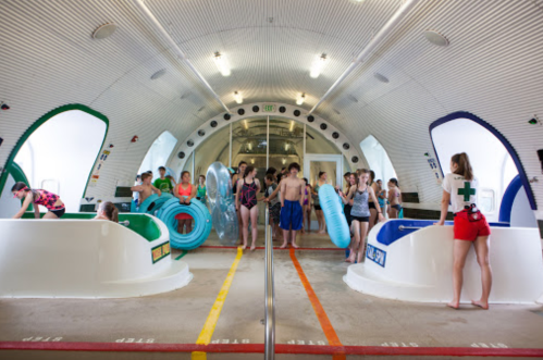 A group of children and teens in swimsuits play in a colorful indoor water park with slides and inflatable rings.
