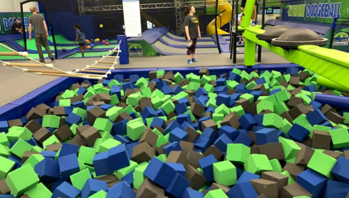A colorful foam pit filled with green, blue, and black cubes, with people playing in a trampoline park.
