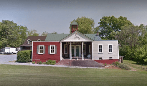 A charming antique shop with a red and white exterior, surrounded by greenery and a gravel parking area.