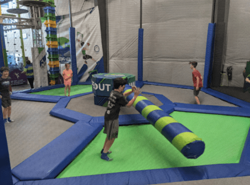 Kids playing on a colorful indoor trampoline park with various obstacles and a foam pit.