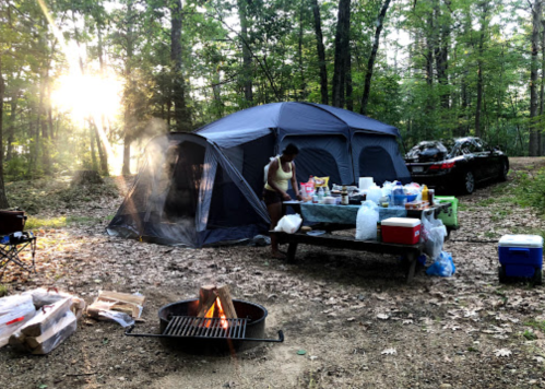 A campsite with a large tent, picnic table filled with food, and a fire pit, surrounded by trees and sunlight.
