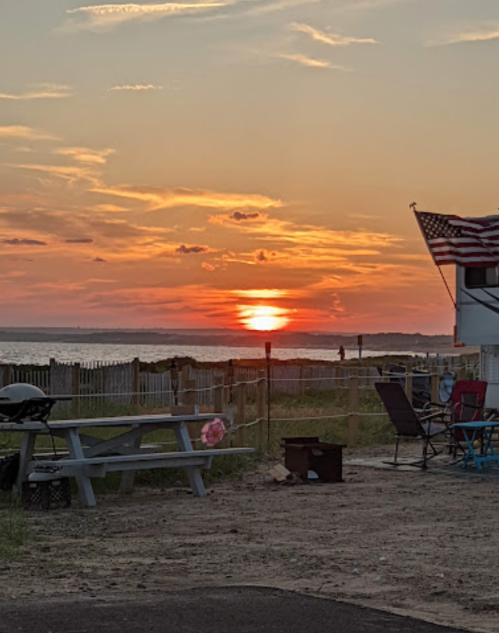 A sunset over the water, with an American flag and picnic table in the foreground, creating a serene outdoor scene.