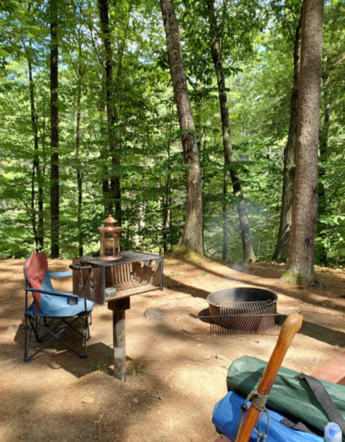 A serene campsite in a forest, featuring a fire pit, a lantern, and two camping chairs surrounded by trees.