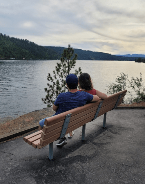 A couple sits on a bench by a lake, surrounded by trees and mountains, enjoying a peaceful view.