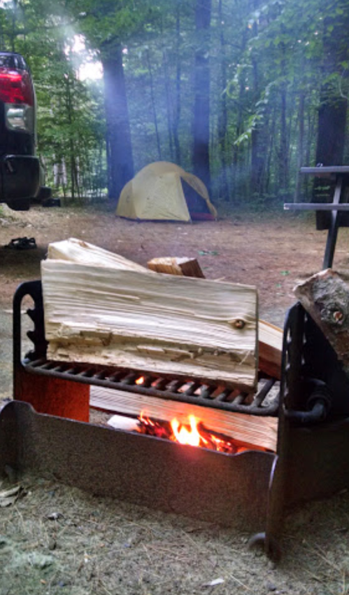 A campfire with logs burning, surrounded by trees, with a tent in the background and smoke rising.