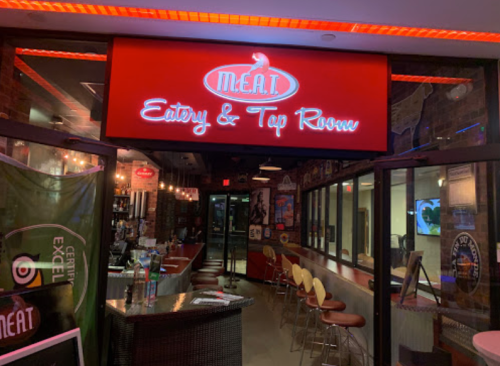 A brightly lit restaurant entrance with a red sign reading "M.E.A.T Eatery & Tap Room" and a modern interior.