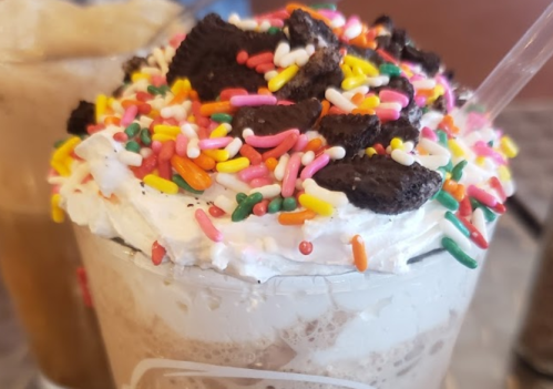 A close-up of a dessert drink topped with whipped cream, colorful sprinkles, and crushed cookies.