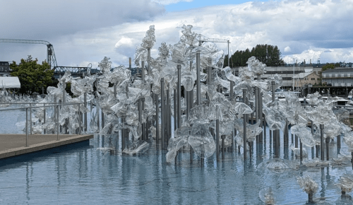 A sculptural installation of clear glass forms rises from a reflective water surface, set against a cloudy sky.