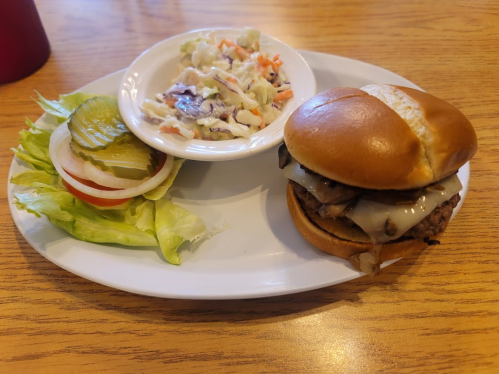 A plate with a burger topped with cheese and onions, a side of coleslaw, lettuce, and pickles.