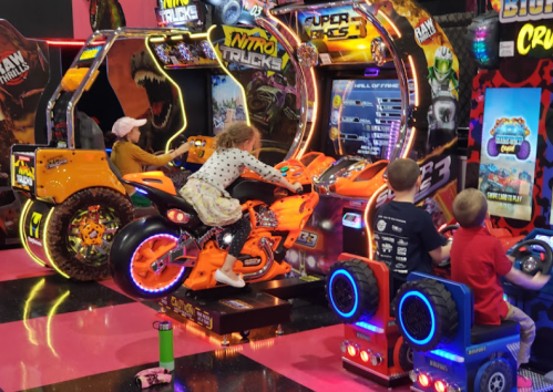 Children playing on colorful arcade games in a vibrant game room filled with bright lights and fun decorations.