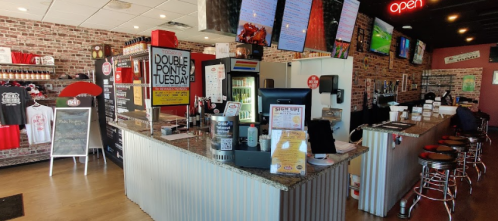 Interior of a casual restaurant with a bar, brick walls, multiple screens, and a counter displaying menus and promotional signs.